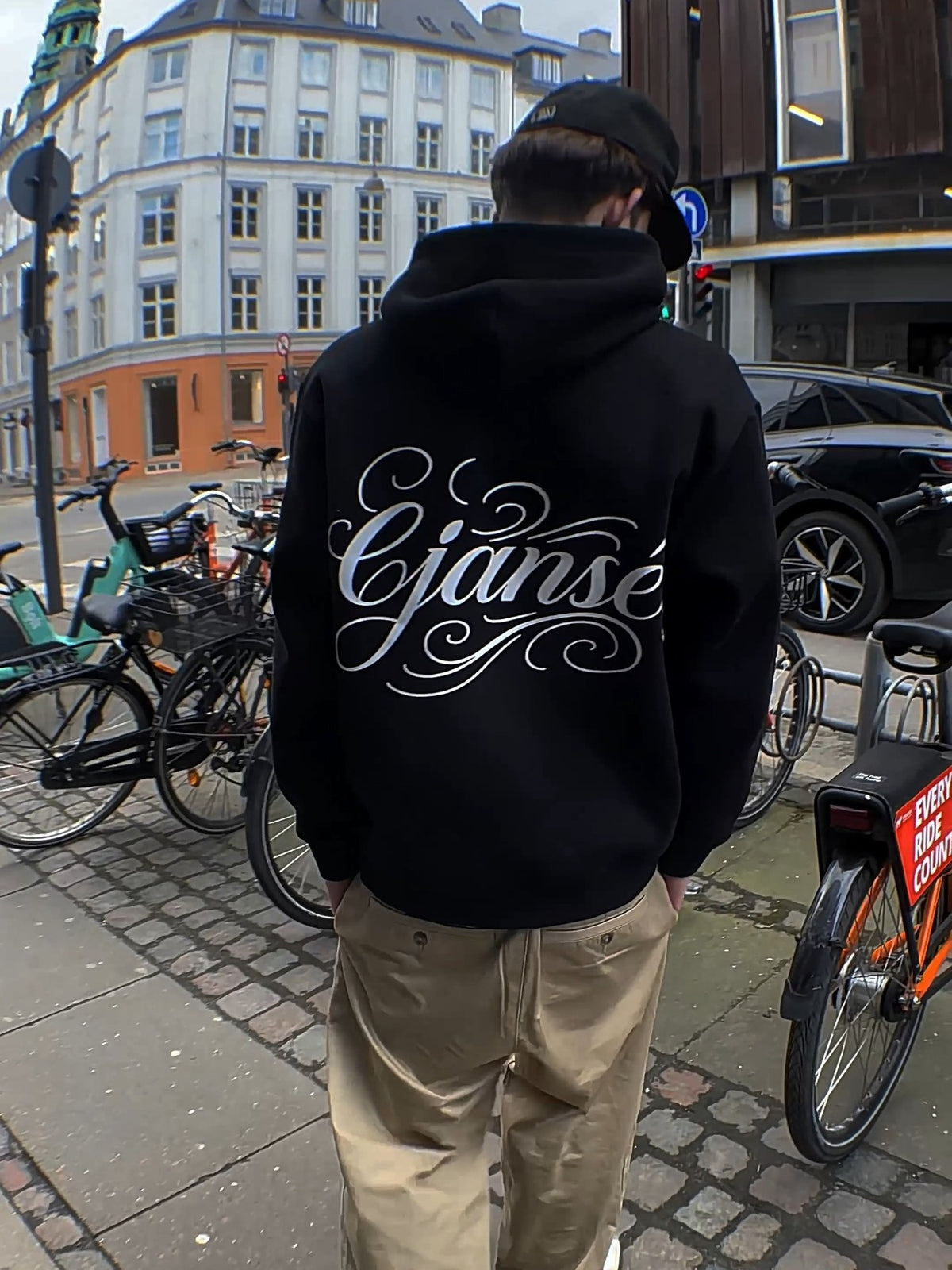 Ryggen af ORIGIN 001, en sort boxy hoodie med stort ornamentalt script-print på ryggen, båret på gaden i København