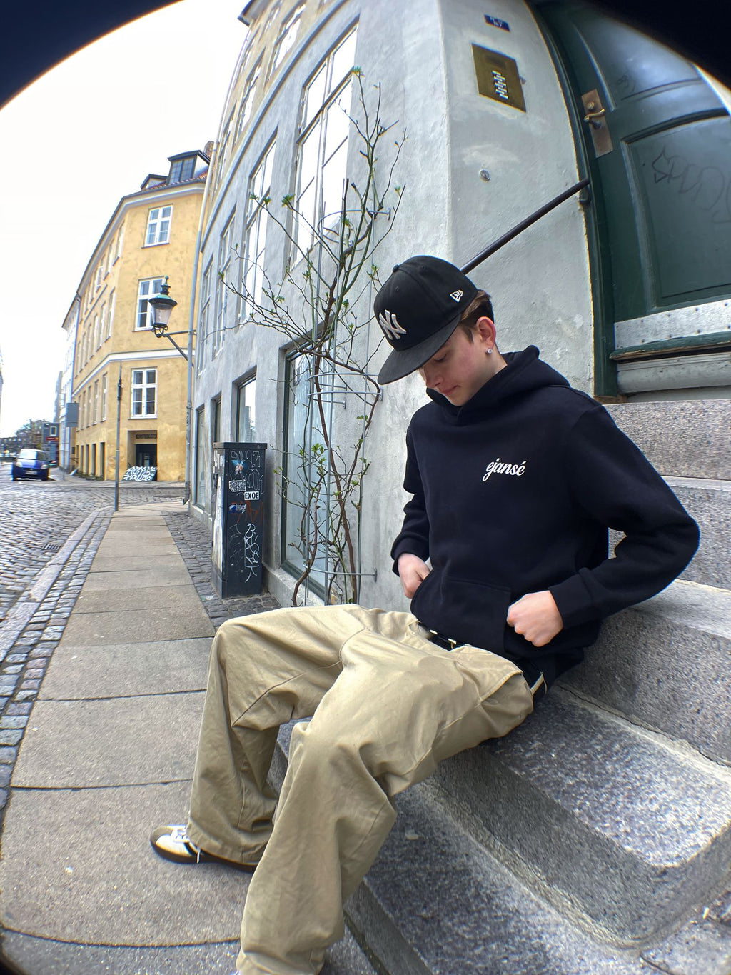 Model sidder på en trappe i sort hoodie fra Ejansé med lille script-logo på brystet