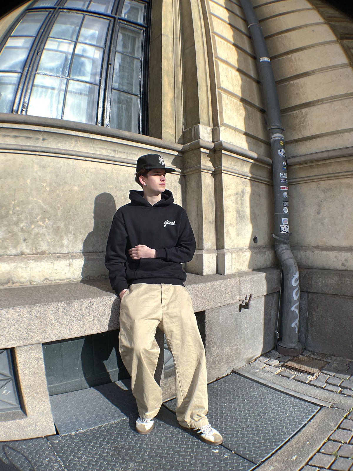 Model i sort boxy hoodie fra Ejansé med lille script-logo på brystet, lænet op ad en facade i København