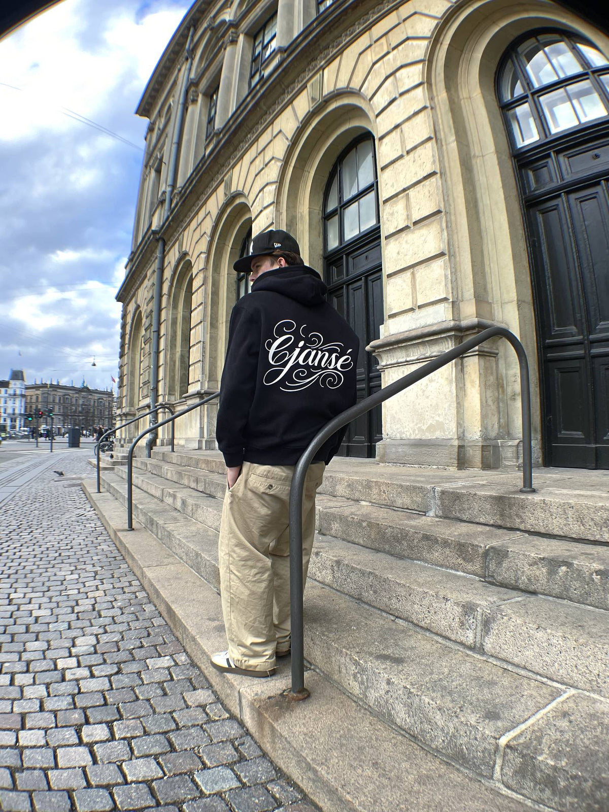Ryggen af ORIGIN 001, en sort hoodie med stort ejanse script-print, båret af model ved en trappe i København